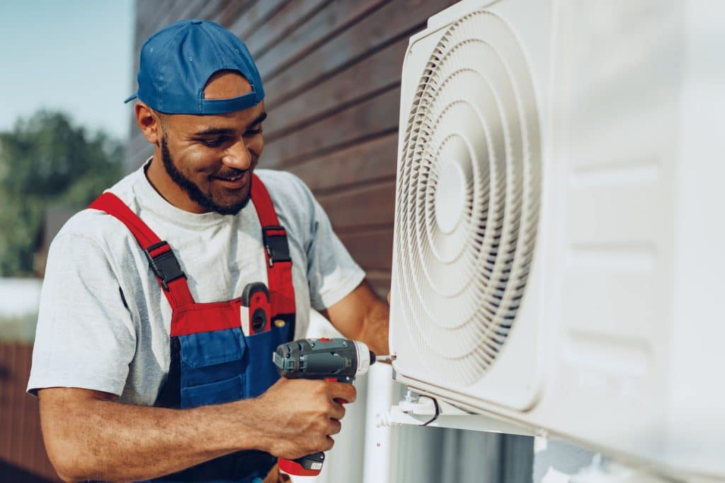 An HVAC technician in protective workwear uses a power drill to install an outdoor air conditioning unit on a building.