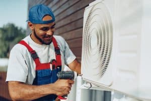 An HVAC technician in protective workwear uses a power drill to install an outdoor air conditioning unit on a building.